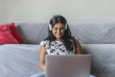 Lächelnde Frau mit Kopfhörern, die zu Hause einen Laptop benutzt - XLGF02330