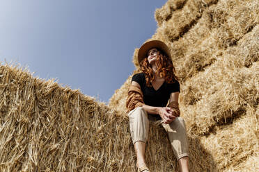 Frau mit Hut sitzt auf einem Heuhaufen auf einem Bauernhof - AFVF09249