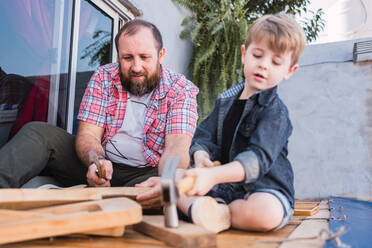 Bearded dad teaching son with hammer working with wood while sitting on boardwalk on weekend - ADSF30982