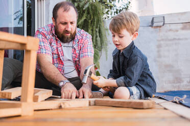 Bärtiger Vater unterrichtet Sohn mit Hammer bei der Arbeit mit Holz, während er am Wochenende auf der Promenade sitzt - ADSF30983