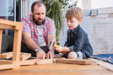 Bearded dad teaching son with hammer working with wood while sitting on boardwalk on weekend - ADSF30983