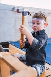 Kind in Jeanshemd und Plastikbrille sitzt mit Hammer gegen Holzstück im Tageslicht - ADSF30991