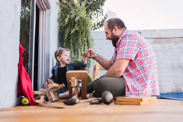 Ground level of cheerful bearded dad in checkered shirt against boy bumping fists while looking at each other against wooden blocks - ADSF30993