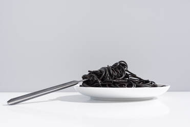 Edelstahl-Gabel in voller Schüssel mit schwarzen Spaghetti mit Tintenfisch Tinte auf weißem Tisch im Studio auf grauem Hintergrund - ADSF31003