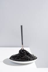 Edelstahl-Gabel in voller Schüssel mit schwarzen Spaghetti mit Tintenfisch Tinte auf weißem Tisch im Studio auf grauem Hintergrund - ADSF31005
