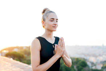 Gelassene Frau mit zusammengelegten Händen auf der Brust und geschlossenen Augen beim Meditieren auf einem Dach während einer abendlichen Yoga-Praxis - ADSF31015