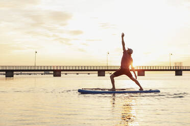 Männlicher Lehrer mit erhobener Hand auf einem Paddleboard im Meer bei Sonnenuntergang stehend - MASF25668