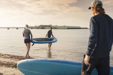 Weibliche und männliche Freunde tragen Paddleboard im Meer am Strand - MASF25732