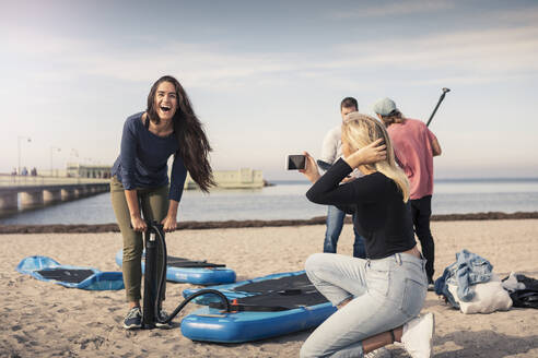 Glückliche Frau, die ihr Paddleboard aufpumpt, während ihre Freundin am Strand fotografiert - MASF25769