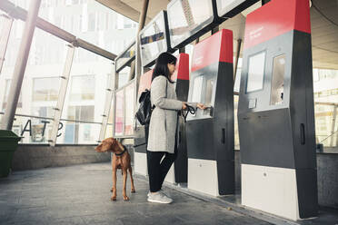 Seitenansicht einer Frau mit Hund an einem Fahrkartenautomaten im Bahnhof - MASF25807