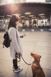 Seitenansicht einer Frau, die ihr Smartphone benutzt, während sie mit ihrem Hund auf dem Gehweg steht - MASF25810