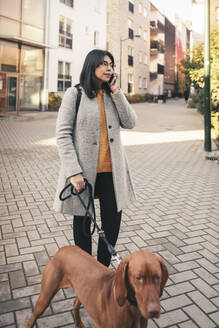 Junge Frau spricht in ihr Smartphone, während sie mit ihrem Hund auf dem Gehweg spazieren geht - MASF25815