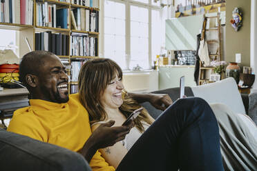 Cheerful couple sitting together with mobile phones on sofa in living room at home - MASF25827