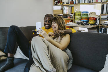 Cheerful man sitting arm around with girlfriend showing smart phone sitting on sofa in living room at home - MASF25830