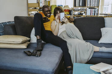 Full length of couple laughing while using smart phone sitting together on sofa in living room at home - MASF25831