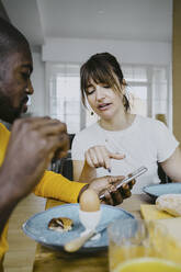 Woman discussing over smart phone with man while having breakfast at home - MASF25836