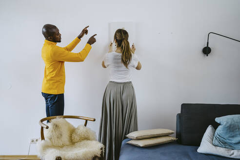 Mid adult man guiding girlfriend adjusting painting on wall at home - MASF25853