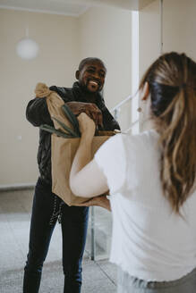 Lächelnder Mann, der einer an der Tür stehenden Frau eine Papiertüte mit Lebensmitteln übergibt - MASF25856