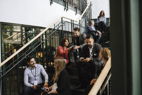 Businessmen and businesswomen discussing while sitting on staircase during networking event - MASF25921