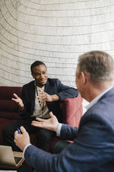 Male colleagues gesturing while discussing together sitting on sofa during networking event - MASF25954