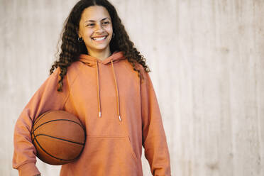 Smiling girl with basketball looking away while standing against wall - MASF26029