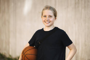 Portrait of smiling girl with basketball against wall - MASF26035