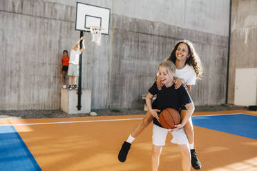 Pre-adolescent girl carrying female friend piggyback at basketball court - MASF26089