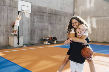 Portrait of girl carrying female friend piggyback at sports court - MASF26090