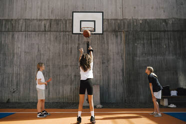 Rear view of girl throwing basketball in hoop while playing with friends at sports court - MASF26115