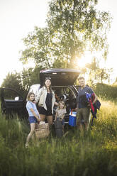 Portrait of mother, father and two daughters standing by car at electric vehicle charging station - MASF26143
