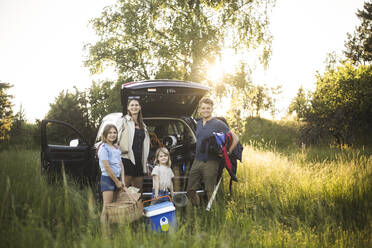 Porträt einer Mutter, eines Vaters und zweier Töchter, die neben einem Auto an einer Ladestation für Elektrofahrzeuge stehen - MASF26147