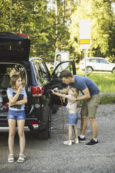 Porträt einer Mutter, eines Vaters und zweier Töchter, die neben einem Auto an einer Ladestation für Elektrofahrzeuge stehen - MASF26163
