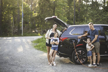 Porträt einer Mutter, eines Vaters und zweier Töchter, die neben einem Auto an einer Ladestation für Elektrofahrzeuge stehen - MASF26164