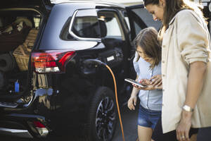 Porträt einer Mutter, eines Vaters und zweier Töchter, die neben einem Auto an einer Ladestation für Elektrofahrzeuge stehen - MASF26175