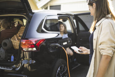 Portrait of mother, father and two daughters standing by car at electric vehicle charging station - MASF26177