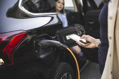 Porträt einer Mutter, eines Vaters und zweier Töchter, die neben einem Auto an einer Ladestation für Elektrofahrzeuge stehen - MASF26179