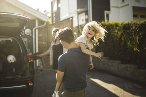 Portrait of mother, father and two daughters standing by car at electric vehicle charging station - MASF26193