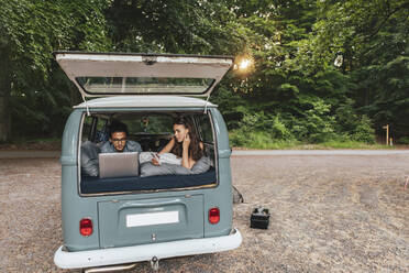 Young man using laptop while female friend with mobile phone lying down in camping van - MASF26233