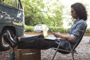 Seitenansicht eines Mannes, der im Urlaub auf einem Stuhl sitzend ein Smartphone benutzt - MASF26247