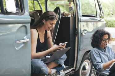 Lächelnde Frau, die ein Smartphone benutzt, während ein männlicher Freund mit einem Buch vor einem Campingwagen sitzt - MASF26249