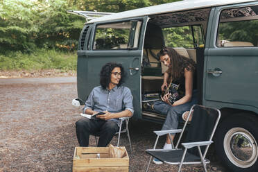 Junge Frau spielt Gitarre, während ein männlicher Freund mit einem Buch neben einem Van sitzt - MASF26259