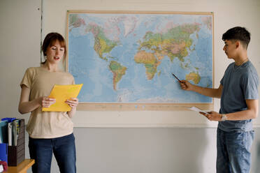 Teenager-Mädchen liest, während ein Junge im Klassenzimmer auf die Weltkarte zeigt - MASF26297
