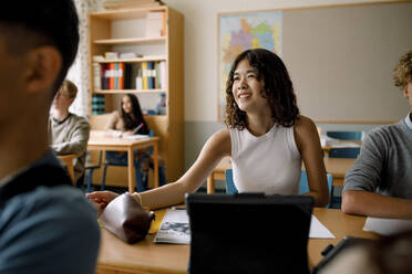 Lächelndes Teenager-Mädchen sitzt am Schreibtisch in der Schule Klassenzimmer - MASF26339