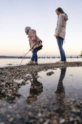 Mädchen spielt mit einem Stock, während sie neben ihrer Mutter am Strand steht - DIGF16548