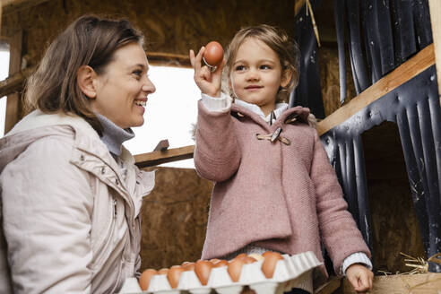 Smiling mid adult woman looking at daughter holding egg - DIGF16554
