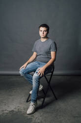 Junger Mann sitzt auf einem Stuhl vor einem grauen Hintergrund im Studio - MEUF04275