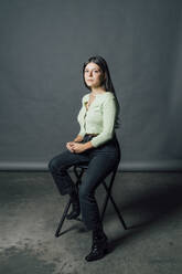 Selbstbewusste junge Frau sitzt auf einem Stuhl im Studio - MEUF04392