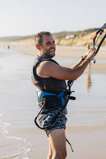 Mann mit Kiteboarding-Ausrüstung am Strand an einem sonnigen Tag - JRVF01974