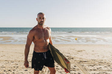 Mann ohne Hemd hält Surfbrett am Strand - JRVF01981