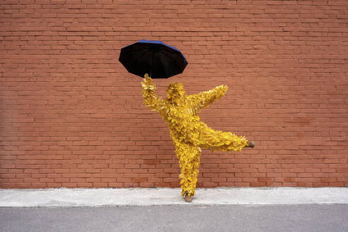 Frau mit Blattkostüm hält Regenschirm beim Tanzen an einer Mauer - VPIF05061